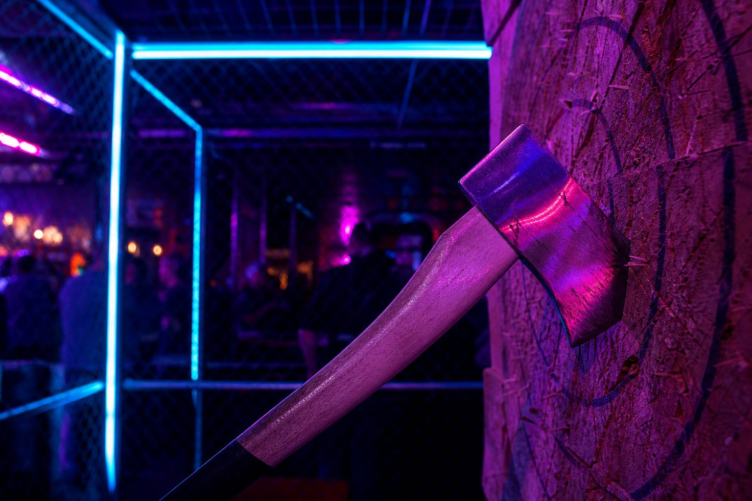 A hatchet buried in an end grain wooden target board inside a chain link throwing cage under pink neon