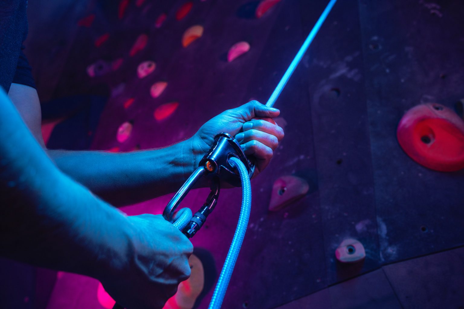 A belayer's hands feeding rope through an assisted braking device in a dim climbing gym lit in cyan