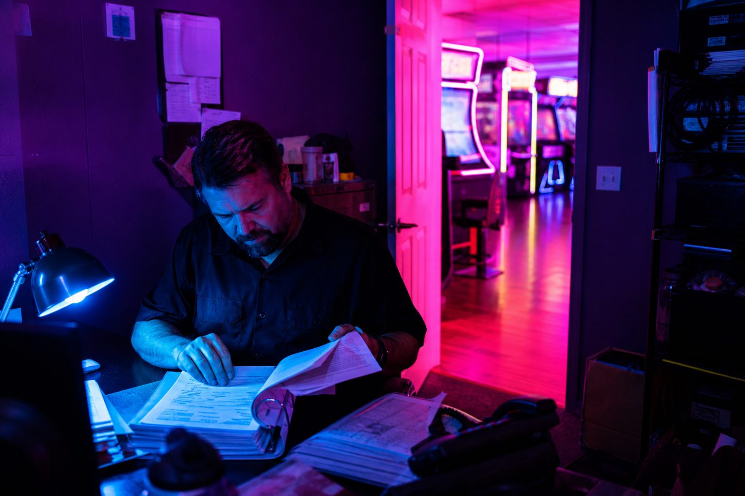 A venue manager reviewing an insurance binder at a back office desk with arcade light glowing behind